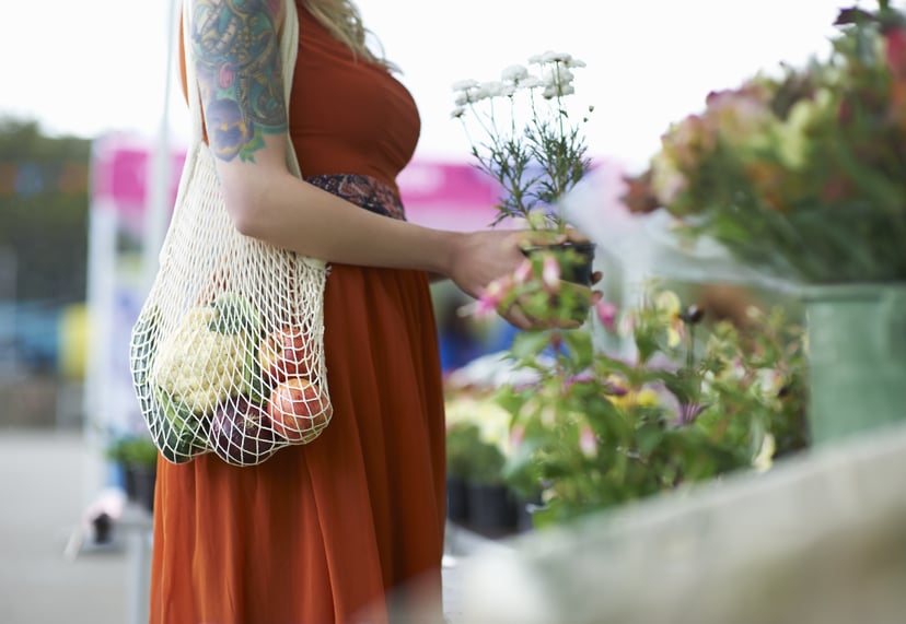 A woman with tattoos shopping on a local market picks up a potted flower plant and carries fresh groceries in her plastic free reusable bag.