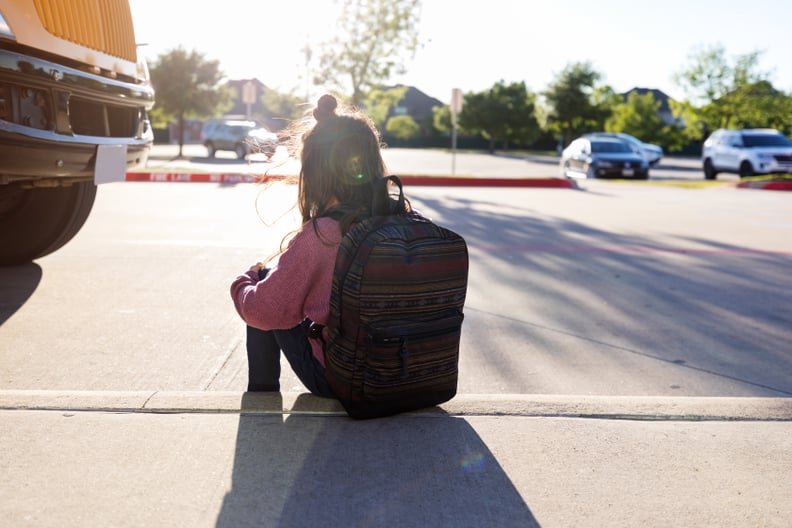 An unknown schoolgirl with a backpack hugs her knees and sits on the curb by herself at a bus stop, while the bus is parked nearby.