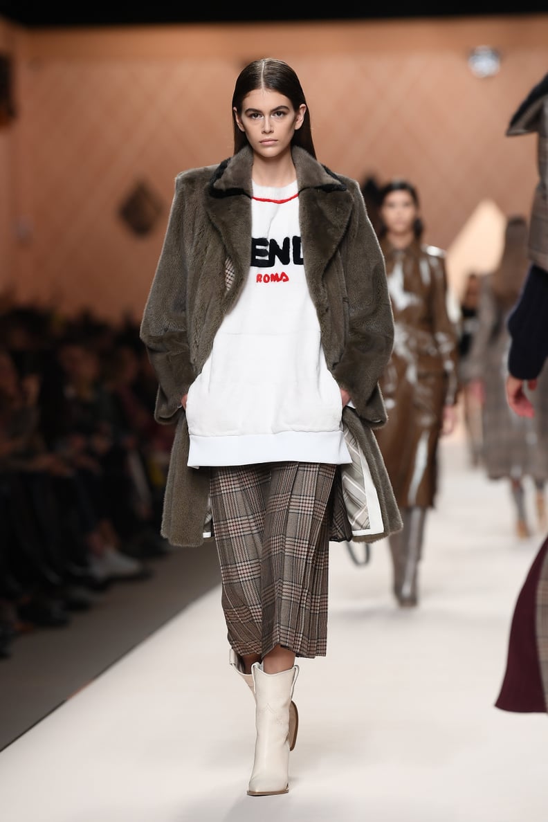 When She Walked For Fendi, Kaia Wore a Logo Sweatshirt With a Plaid Skirt, Fur Coat, and White Ankle Boots