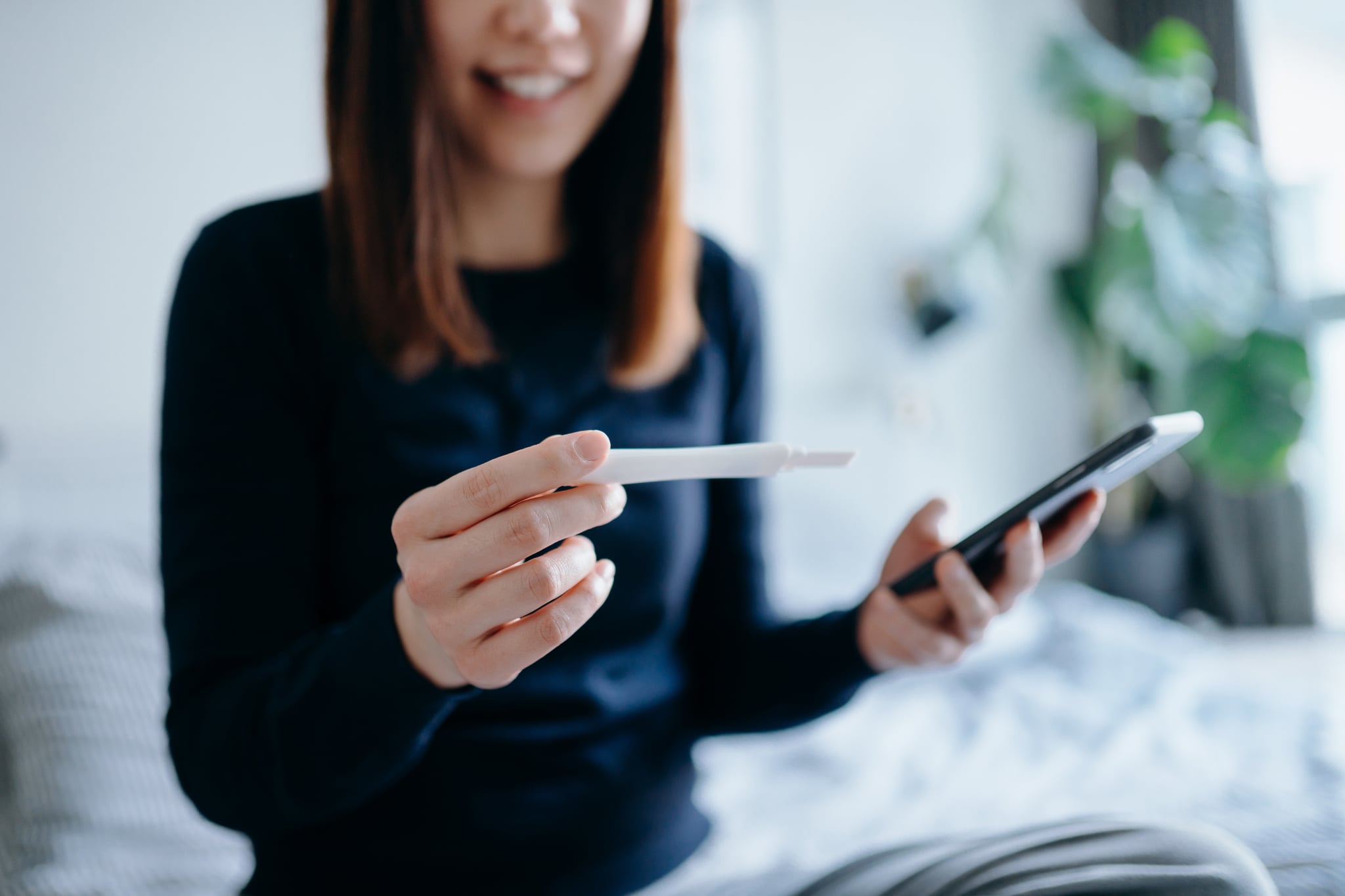 Joyful young Asian woman smiling happily, using pregnancy tracker apps on smartphone to check the pregnancy test result. It's finally happening. The long-awaited news. Life events, fertility and family concept