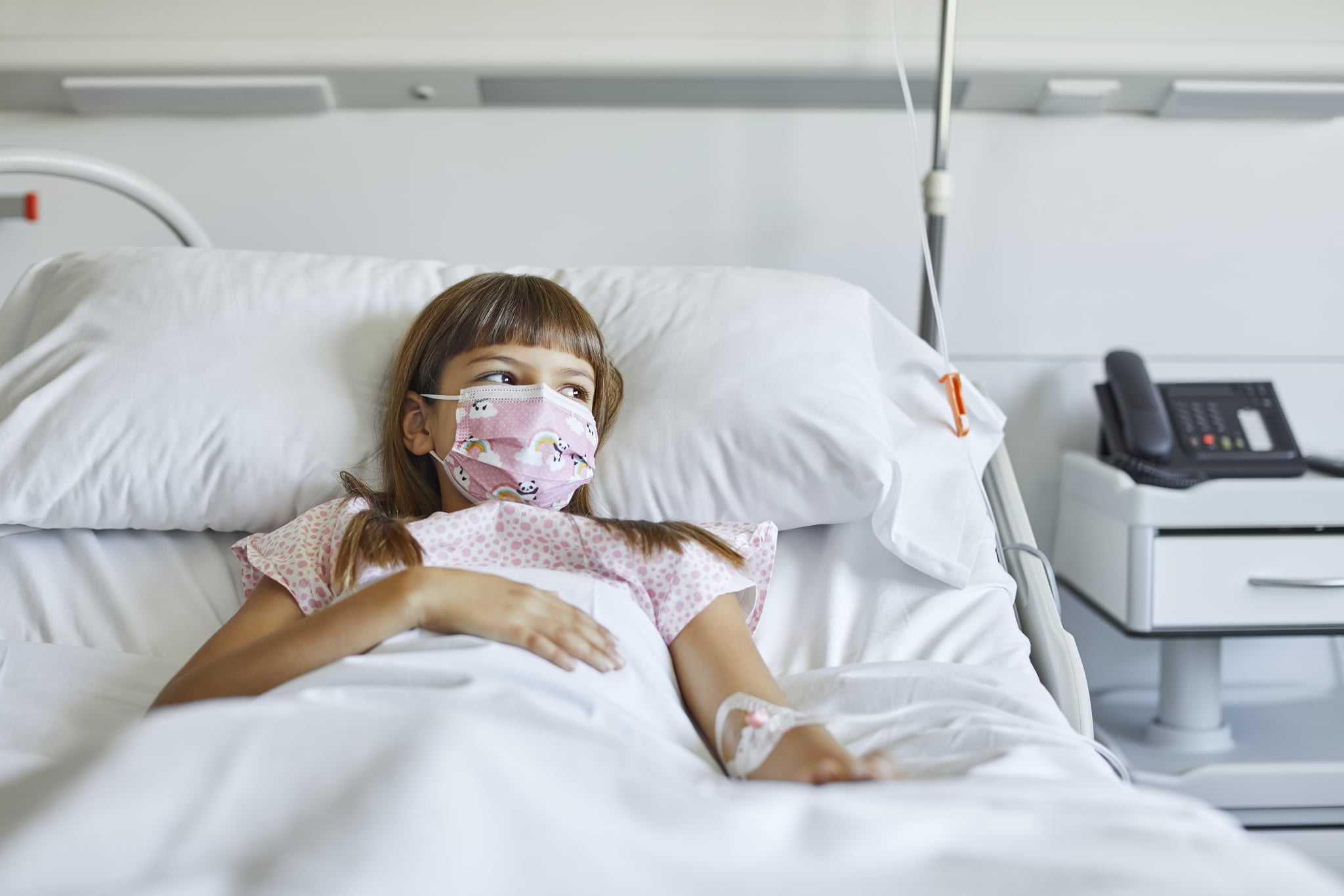 Sick girl lying on bed in ICU. Female patient is looking away while wearing protective face mask. She is in hospital during COVID-19 pandemic.