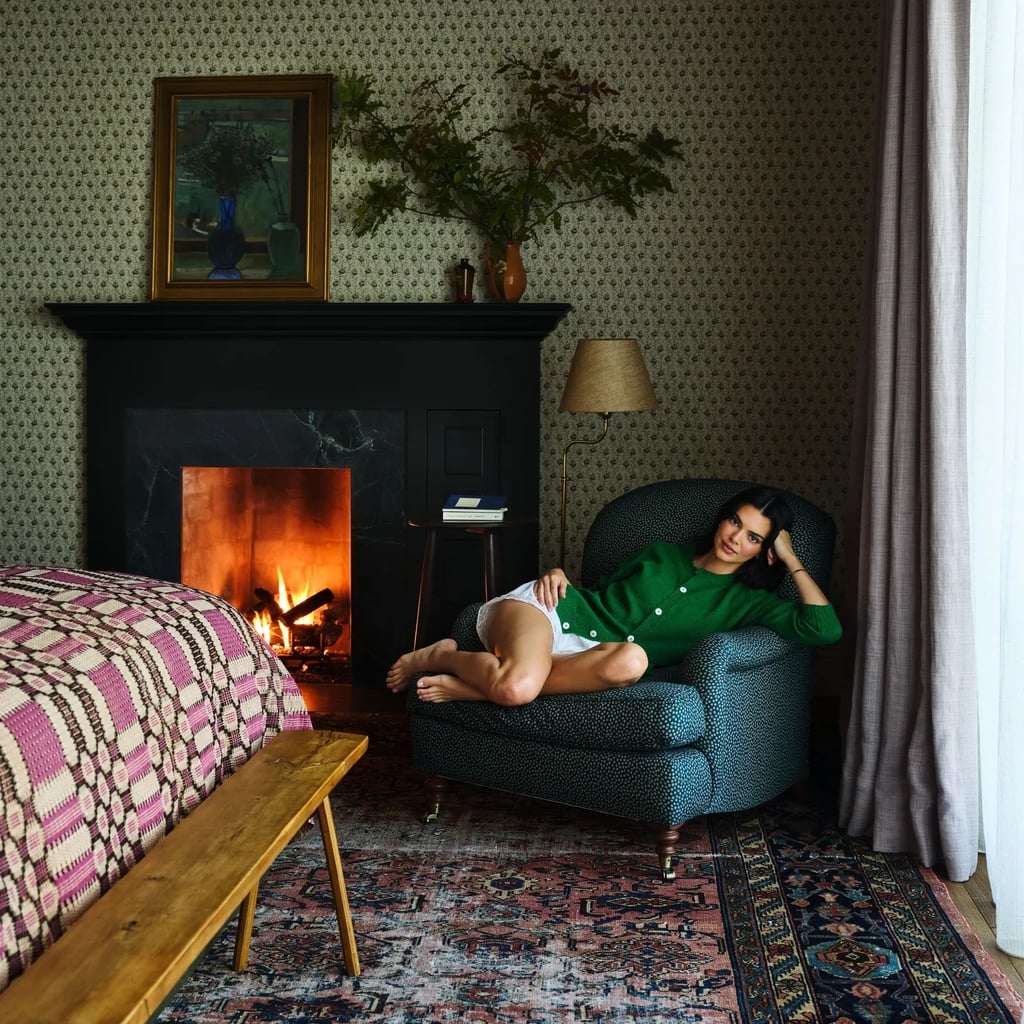 Kendall Jenner lounges on a patterned armchair beside a lit fireplace, wearing a green cardigan and white shorts, looking relaxed in a cozy, vintage-style bedroom.