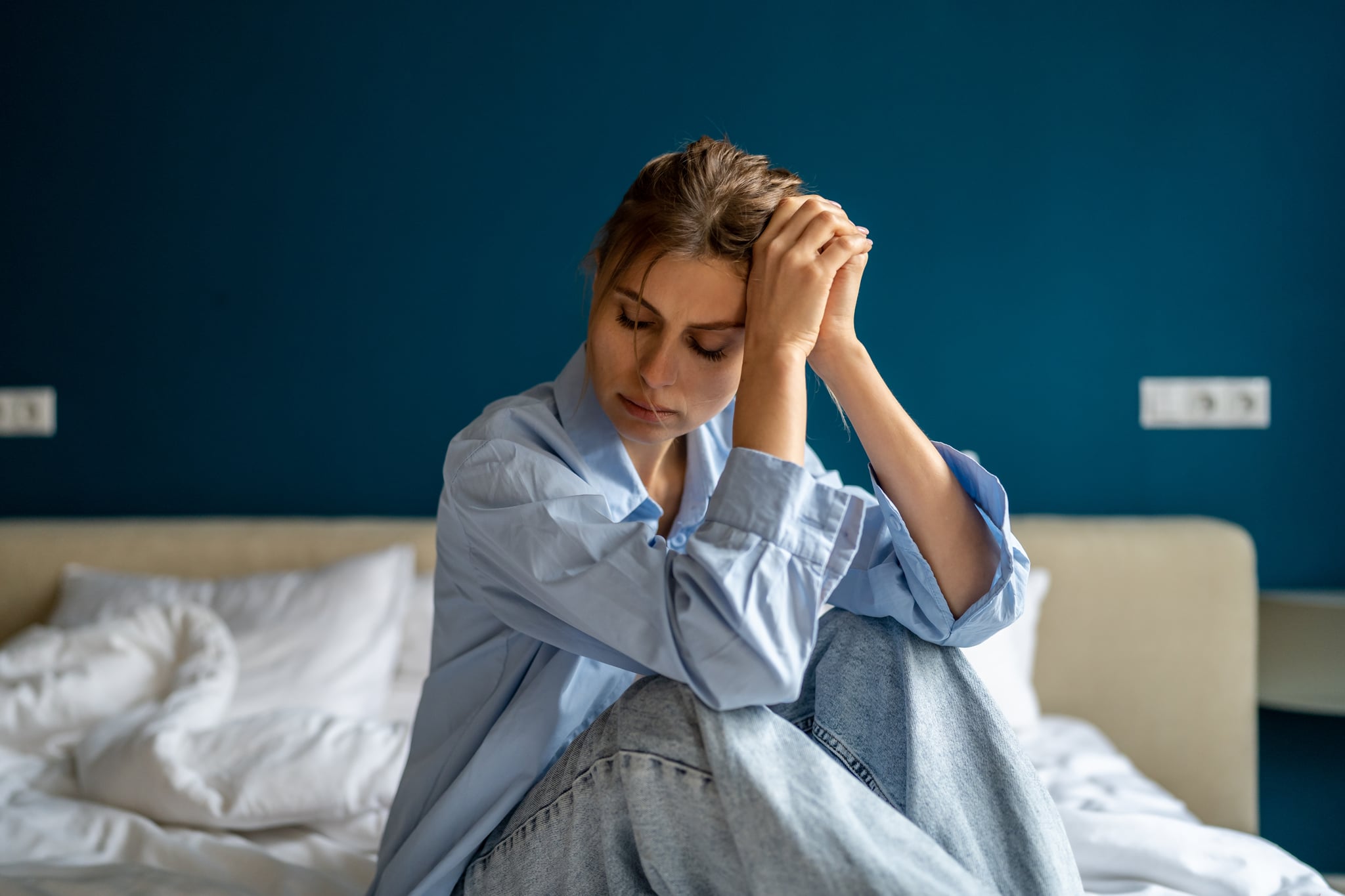 Young fair-skinned unhappy woman sitting on bed at home wearing blue button-up and light wash jeans.