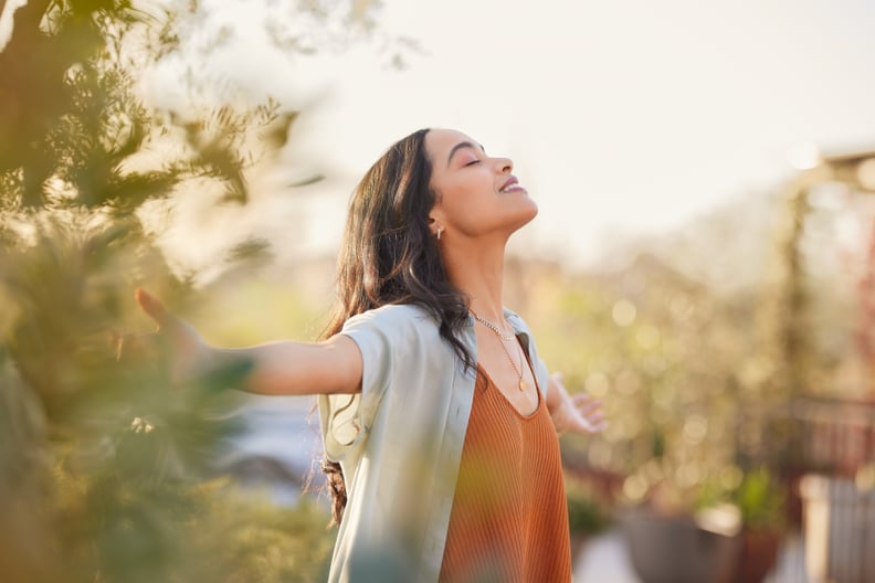 Young latin woman with arms outstretched breathing in fresh air during sunrise at the balcony. Healthy girl enjoying nature while meditating during morning with open arms and closed eyes. Mindful woman enjoying morning ritual while relaxing in outdoor par
