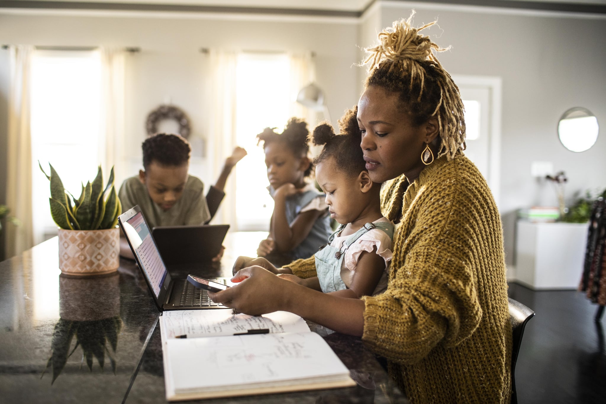 Mother working from home while holding toddler, family in background.