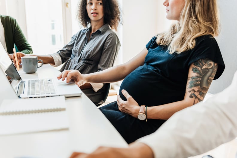 A pregnant woman sitting and working on a laptop alongside some of her colleagues at the office.