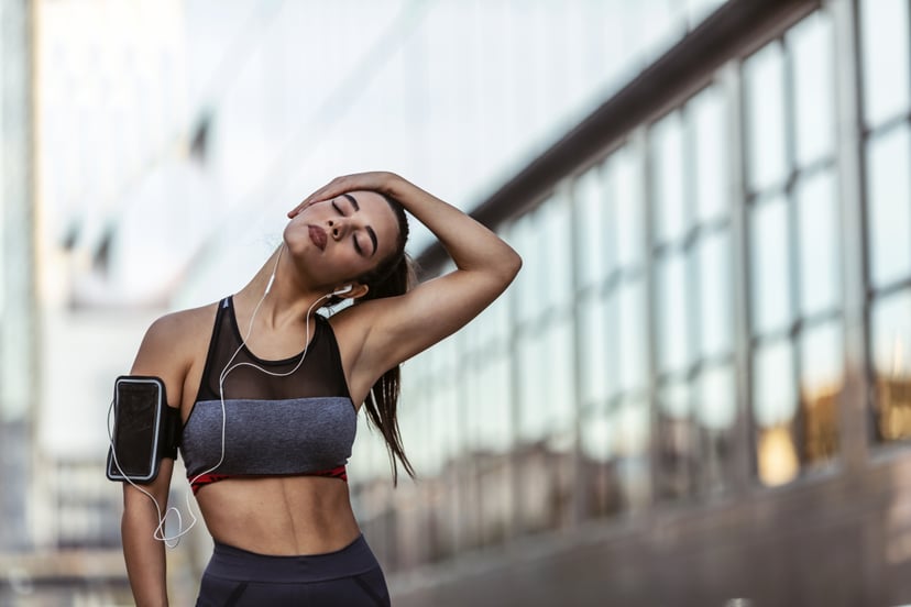 Outdoor shot of beautiful dark hair Caucasian woman athlete wearing black, gray sports bra warming up neck before workout, standing against city with copy space for your promotional content.