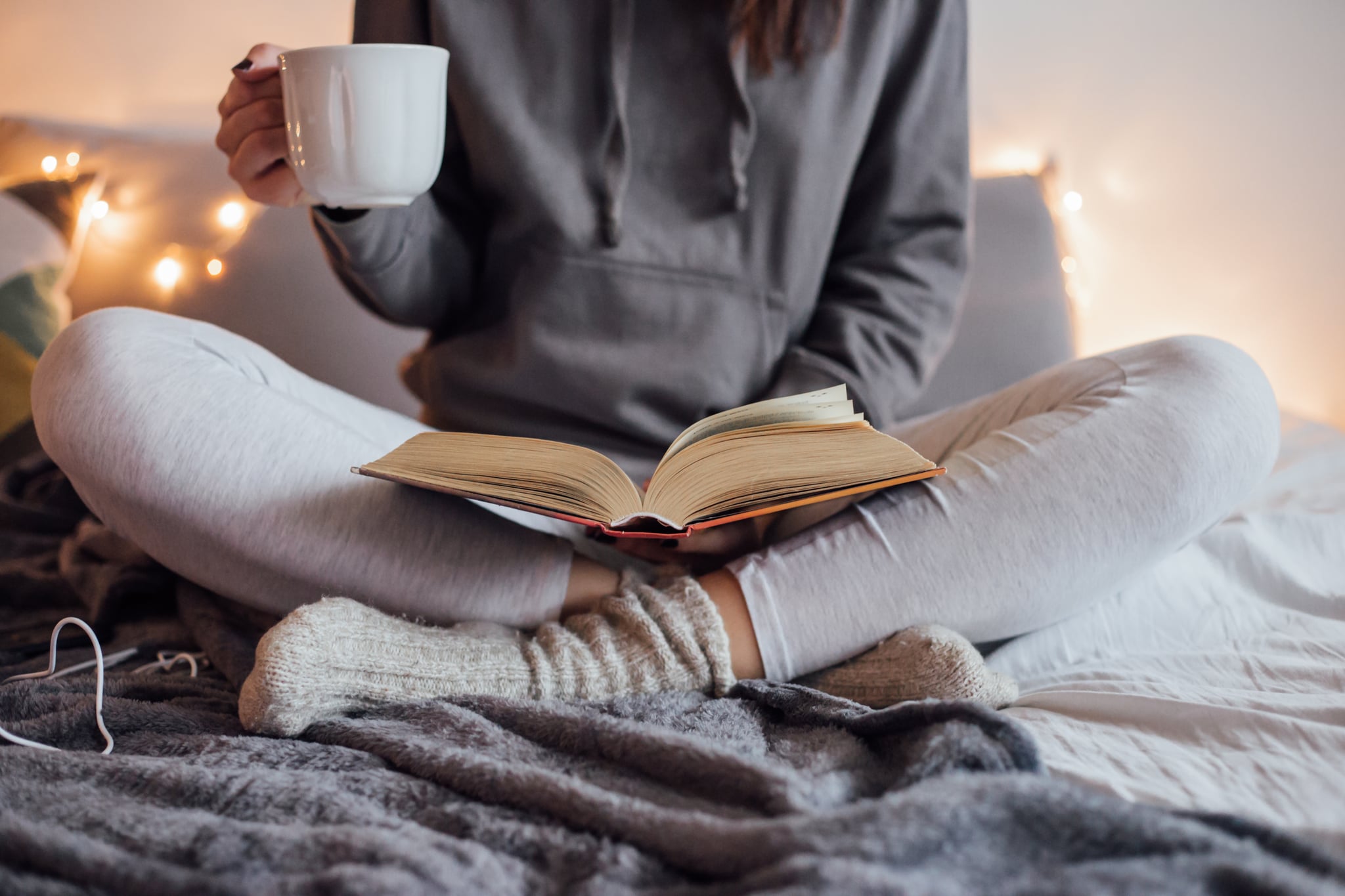 Girl holding cup of hot tea and reading in bed. Around her in bad earphones, book, smart phone. Decorative lights in background.