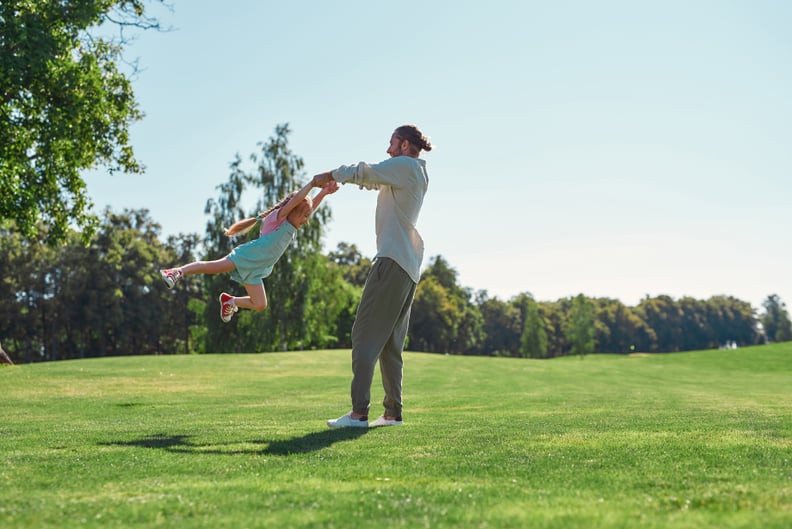 Full length shot of young father and daughter having fun, spinning together on grass field in summer park. Childhood, parenting, happiness concept
