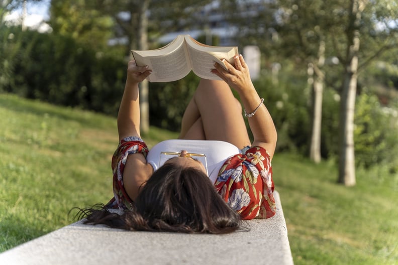 Woman enjoying reading a book while relaxing lying on a bench in a park.