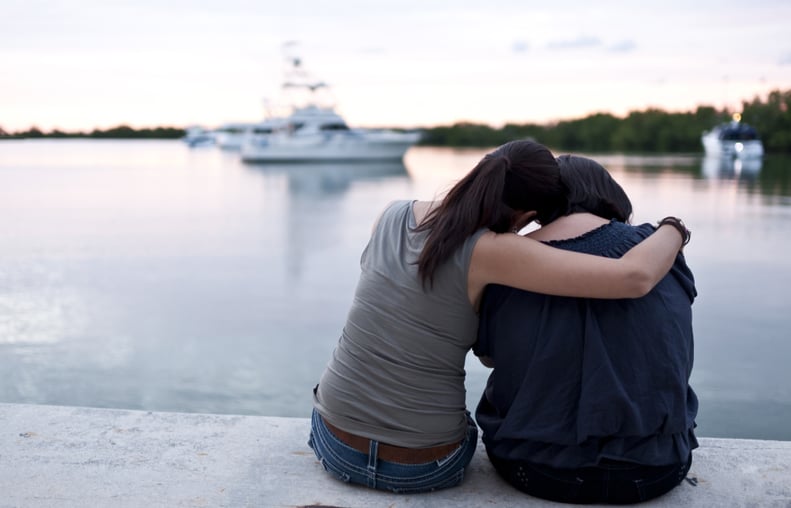 Two mature hispanic women sitting at the docks embracing from behind