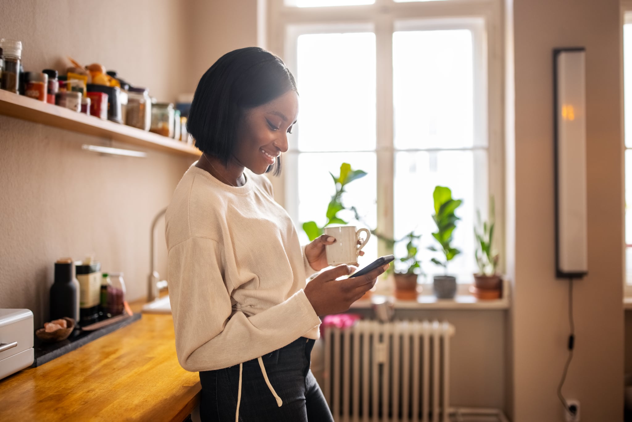 Woman standing in the kitchen holding a cup of coffee and texting on her cell phone. Woman using a mobile phone at home.