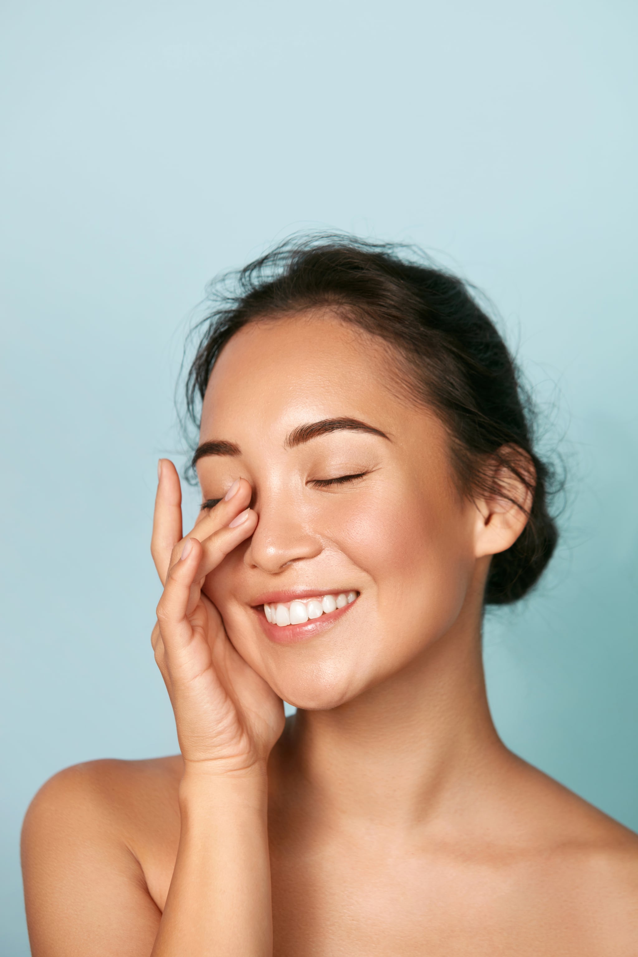 Beauty face. Smiling asian woman touching healthy skin portrait. Beautiful happy girl model with fresh glowing hydrated facial skin and natural makeup on blue background at studio. Skin care concept