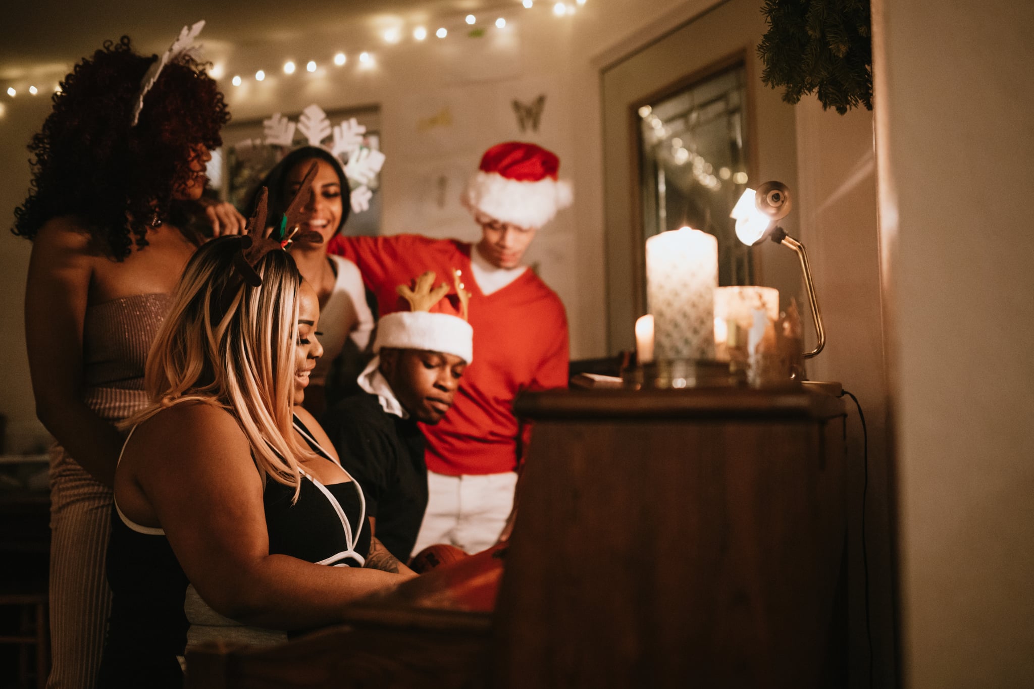 A group of young adult friends gather at a home for Christmas celebration over the holiday, dressed to fit the occasion with various Christmas accessories.  They sing songs together at the piano, enjoying the Christmas cheer.