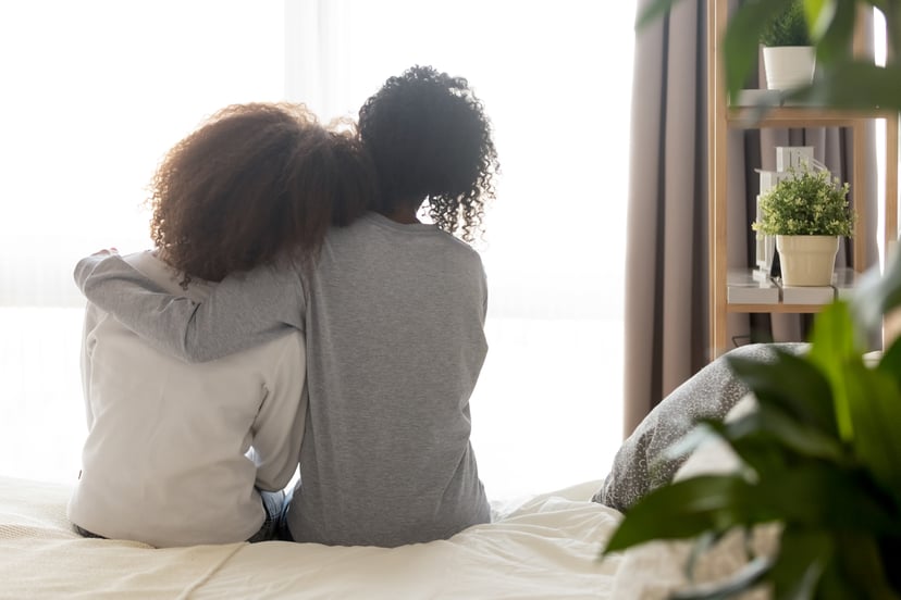 Loving african american single mother sister embrace teen daughter sit on bed looking at window, parent mom hug support protect teenage girl, family trust hope talk understanding concept, rear view