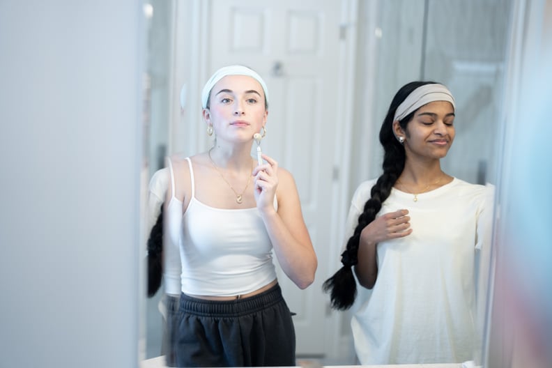A small group of teen girls are seen in a bathroom, standing in front of a mirror as they each tackle their skin care routines.  The friends are dressed comfortably and are focused on the task.