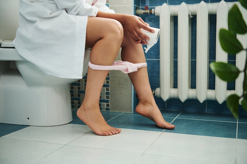 Woman in bathroom sitting on toilet with toilet paper in hand and panties pulled down.