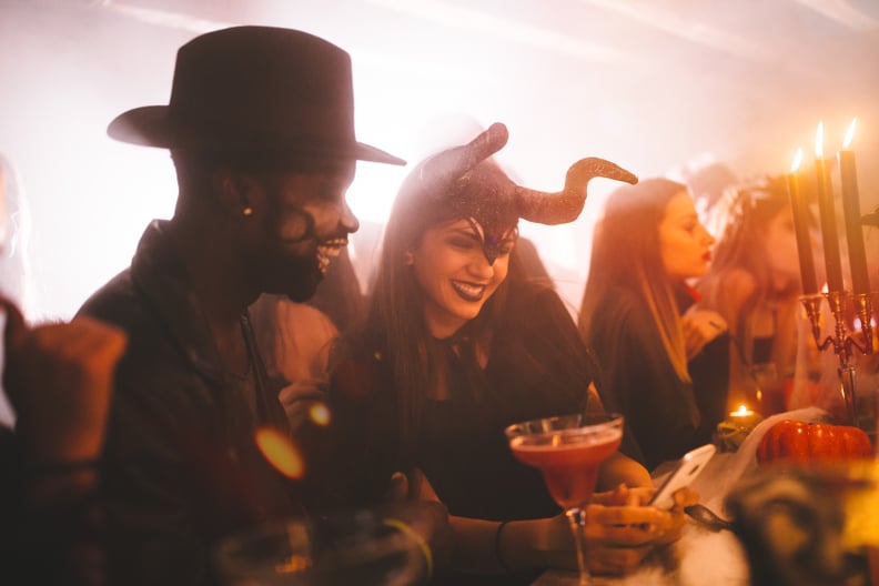 Young multi-ethnic couple in scary costumes having fun using smartphone and texting at Halloween party