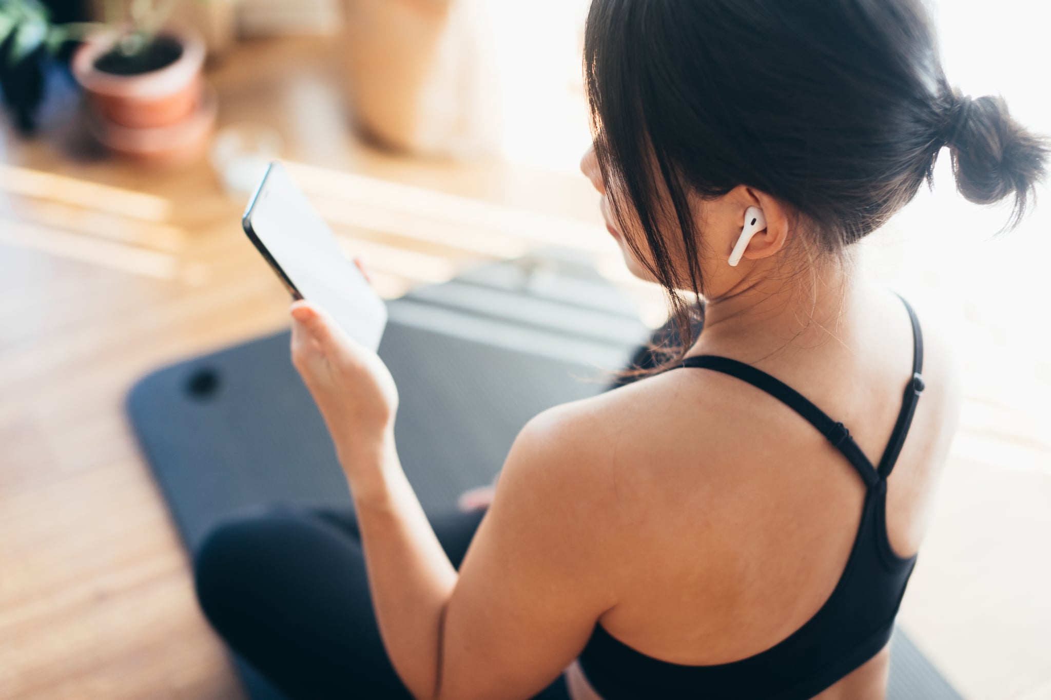Rear View of beautiful young Asian woman listening music with earphone and checking social media on smartphone, sitting on the exercise mat after practicing yoga.