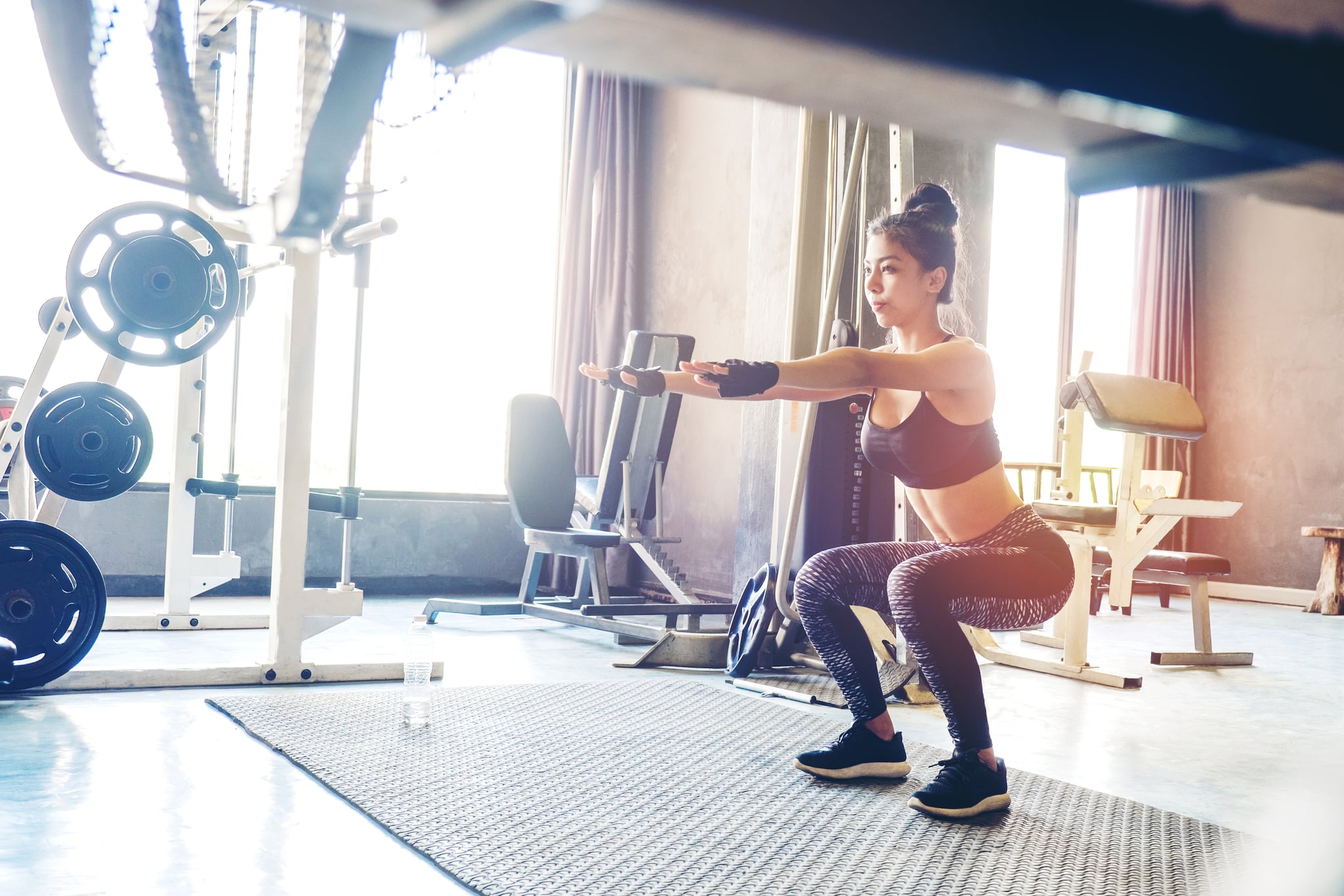 Deep squat of young beautiful woman in sportswear doing squat while standing in front of glass at gym