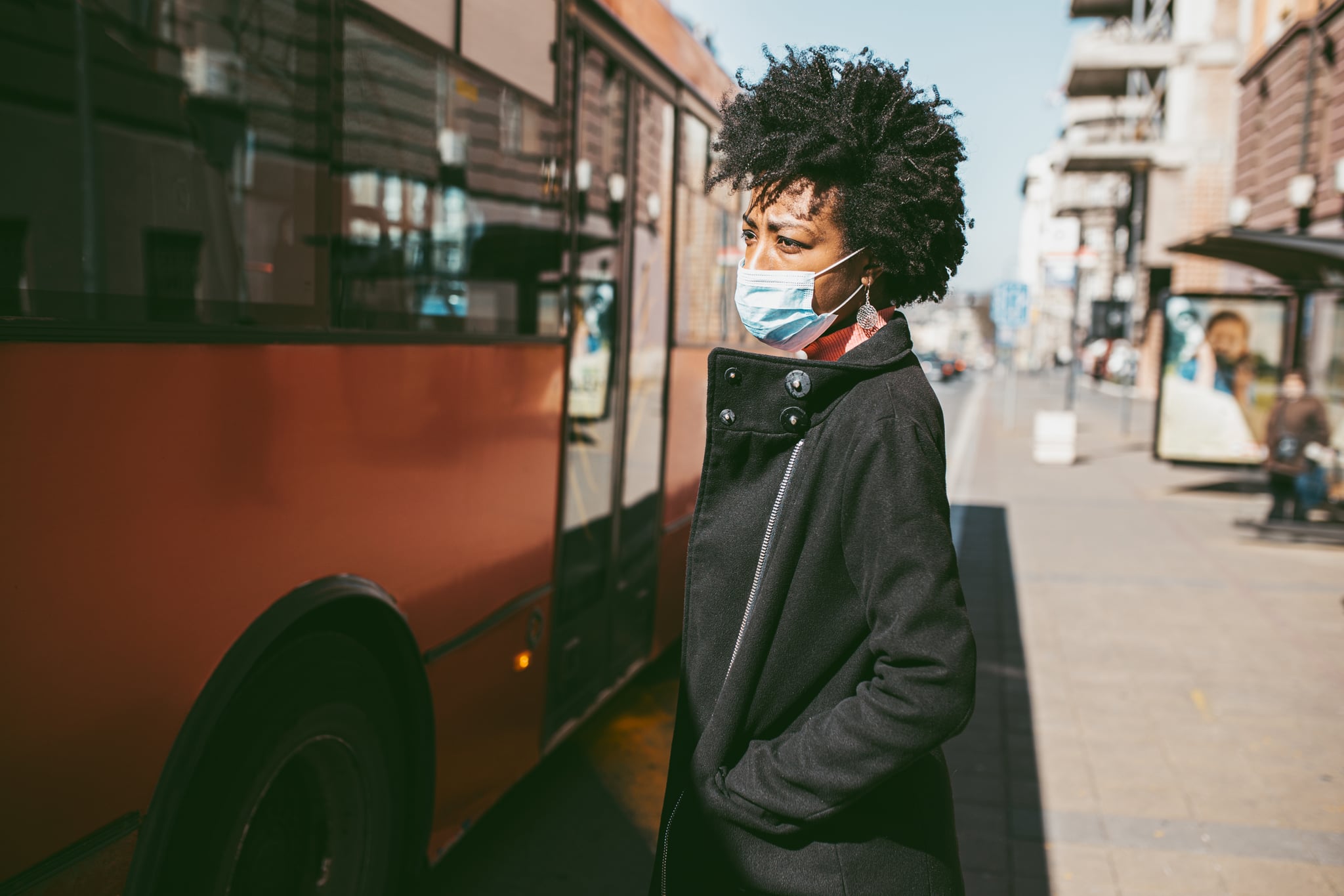 Young Afro American woman standing on city street with protective mask on her face and waiting for bus transportation. Virus pandemic and pollution concept.