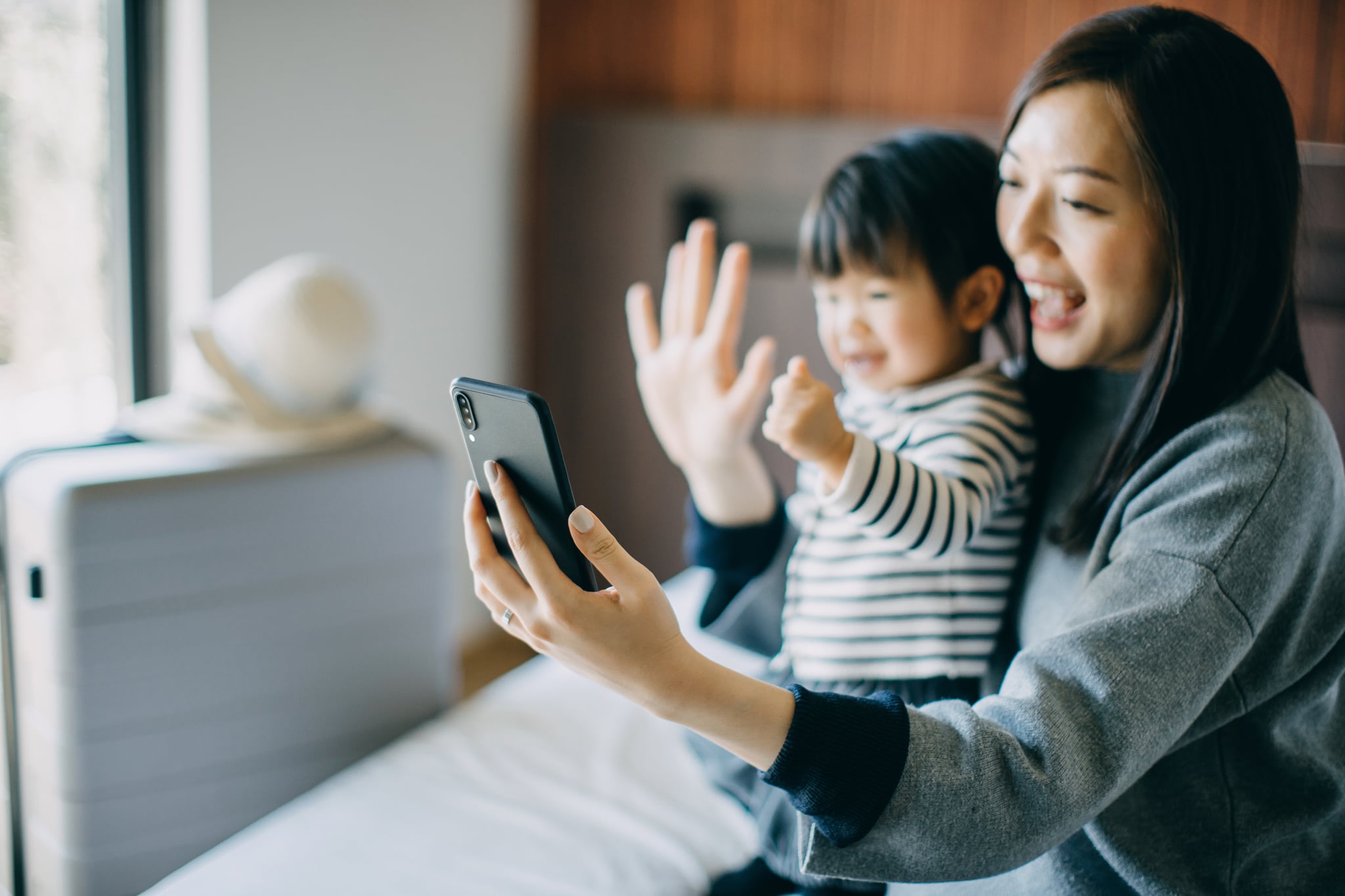 Young Asian mother and cute little daughter having video call on smartphone with family in hotel room while on holiday and smiling joyfully