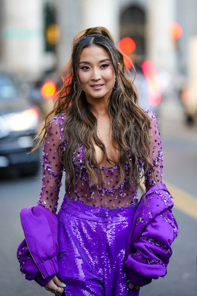 NEW YORK, NEW YORK - FEBRUARY 11: Ashley Park wears silver earrings, a gold chain necklace, a pale purple tulle with embroidered rhinestones / crystal V-neck / long sleeves body, a purple nylon with embroidered crystal / rhinestones striped print pattern 