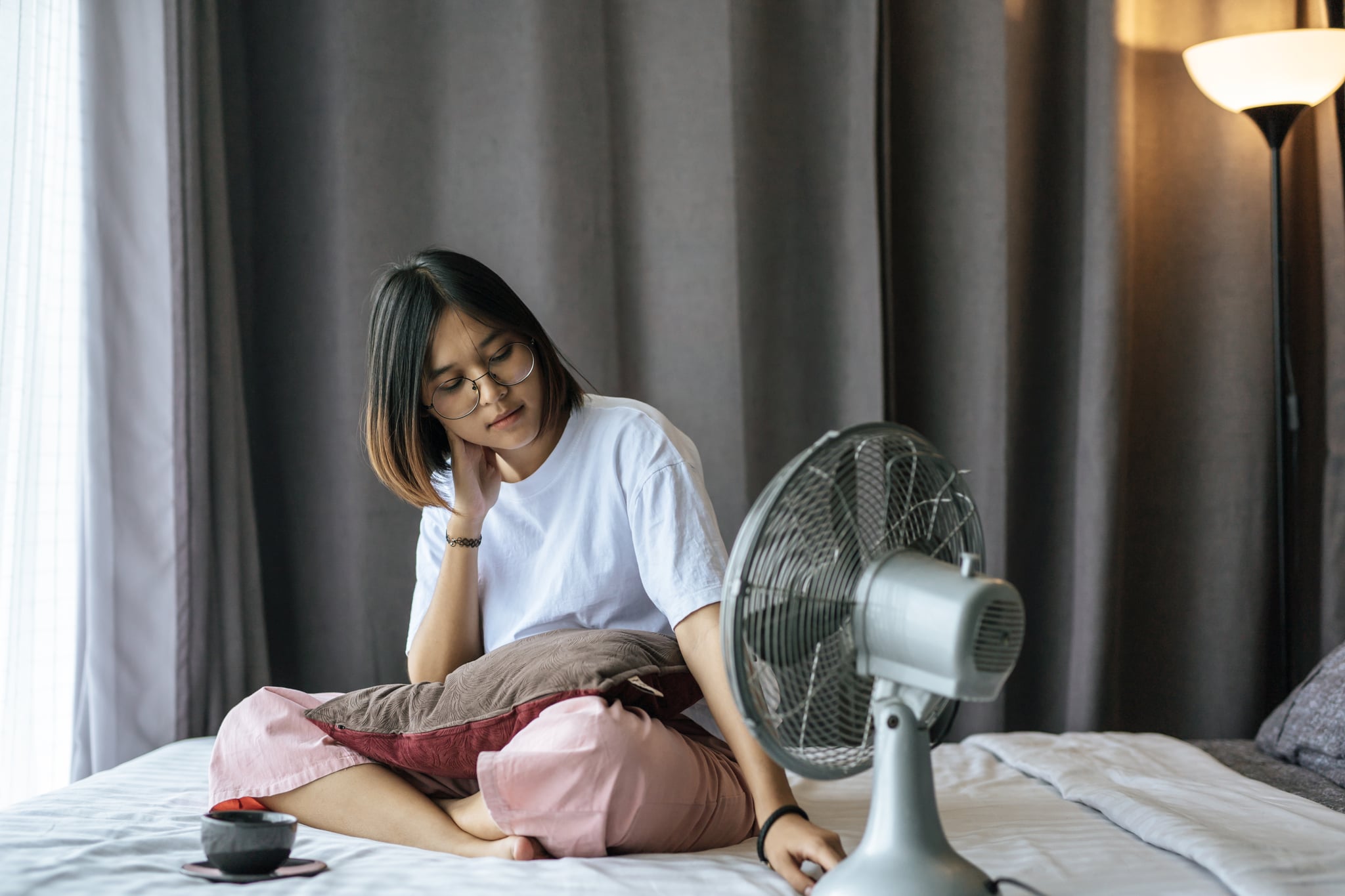 There was a girl sitting and drinking coffee in the bedroom and a pillow on her legs.