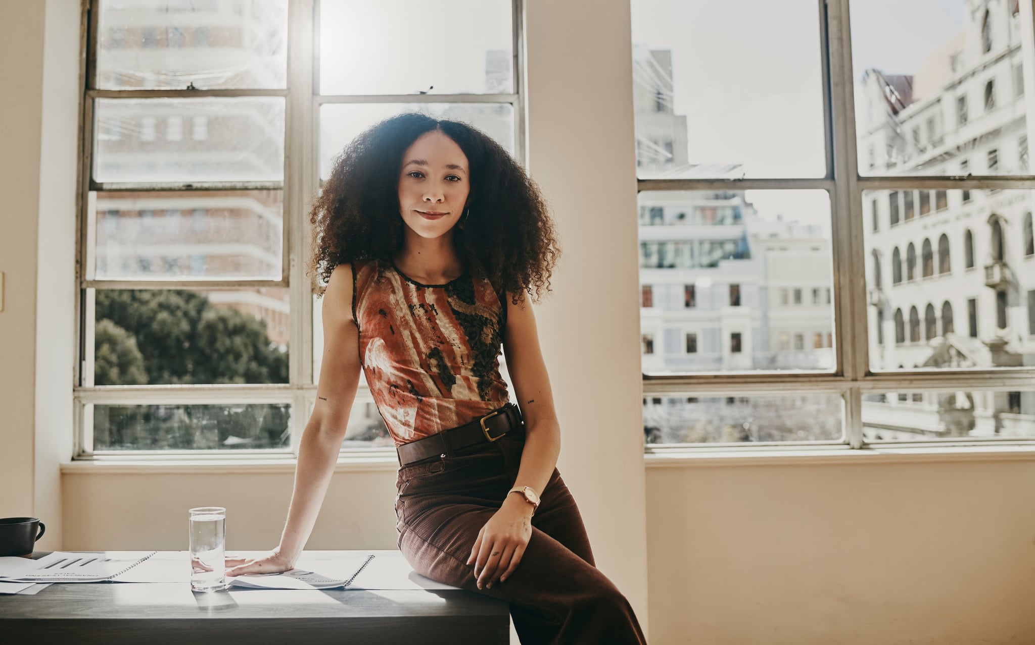 Woman in office sitting on desk and looking at camera