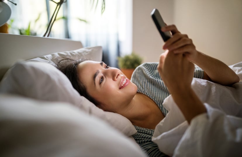 Young woman lying in her bed and using smart phone. Woman waking up in her bed and checking her phone.