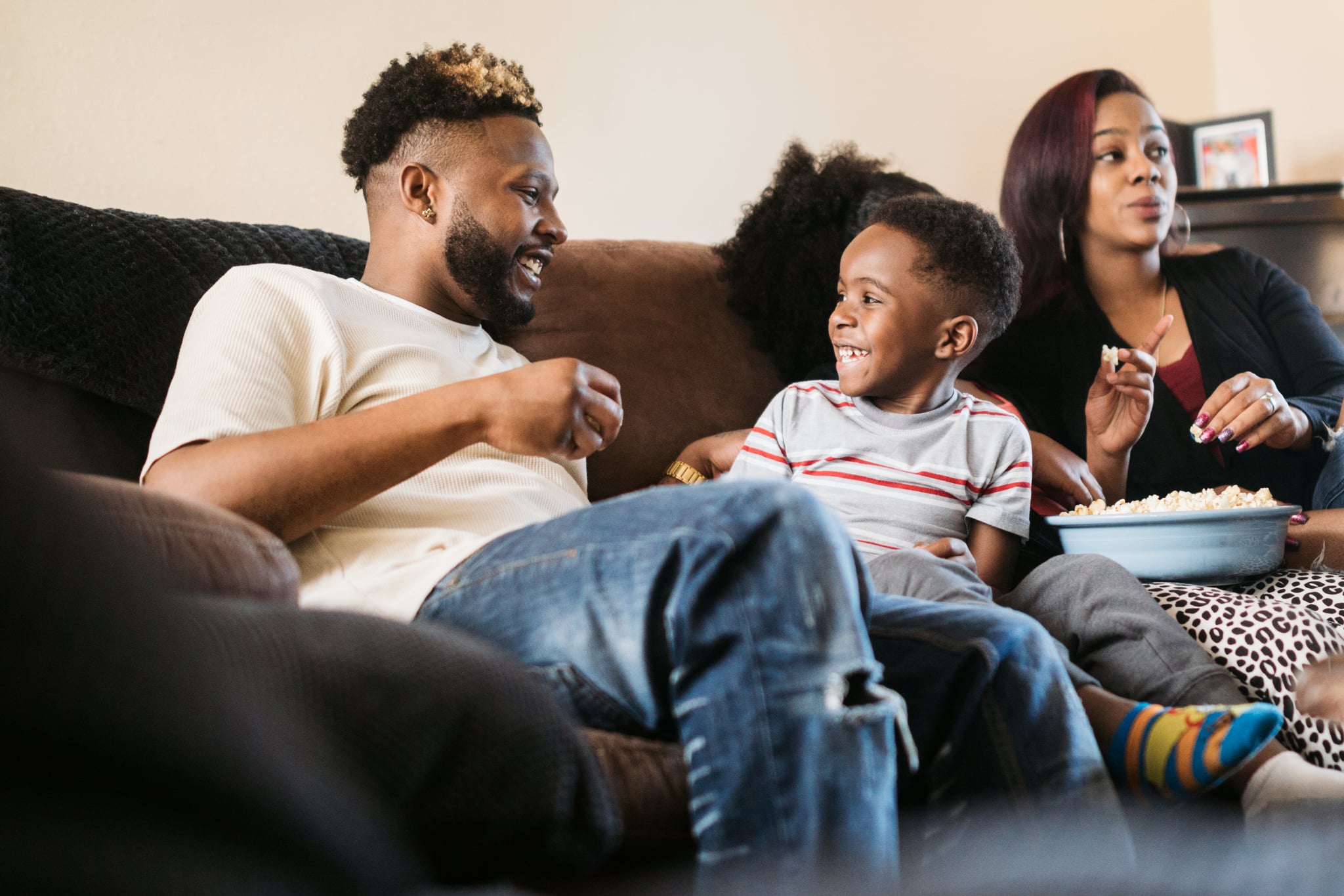 A fun African American family gets cozy on the sofa with a bowl of popcorn for their weekly movie tradition.  A time of connection and tenderness.