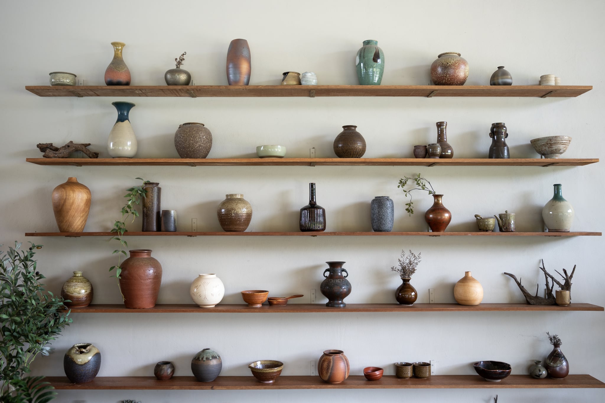 A straight-on, full-frame shot of a wall with five wooden shelves displaying a diverse collection of ceramic vases and bowls. The pieces vary in shape, size, color, and texture, creating a visually rich and eclectic display. The background is a simple, light-colored wall, allowing the focus to remain on the pottery. This image is ideal for use in design, home decor, or art-related projects. The variety of ceramic pieces and the clean, organized presentation contribute to a visually appealing composition.