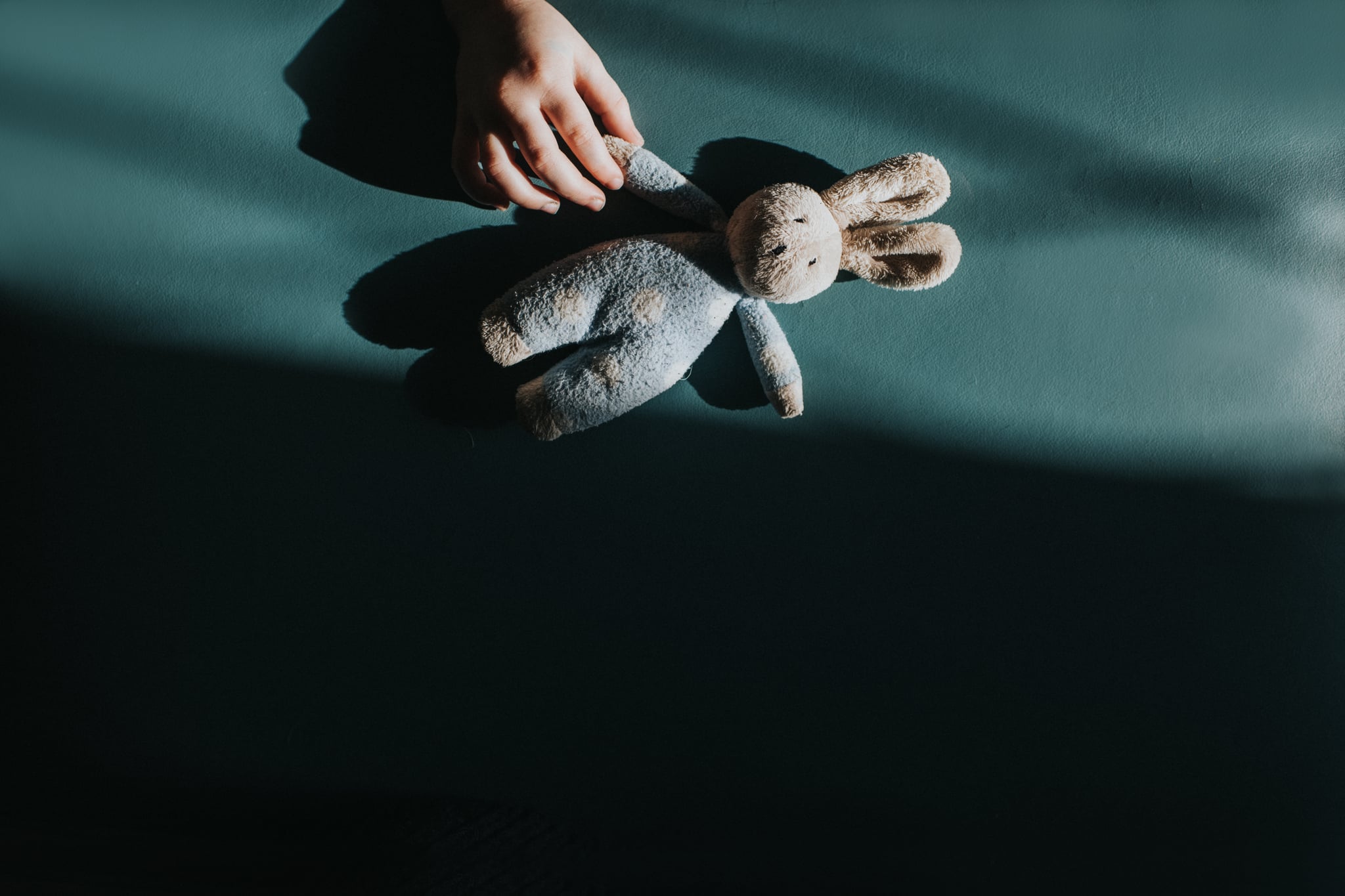 Conceptual image of a small child's hand reaching to grab a small soft blue toy bunny by the paw. The toy is illuminated by sunlight and shadows provide space for copy.