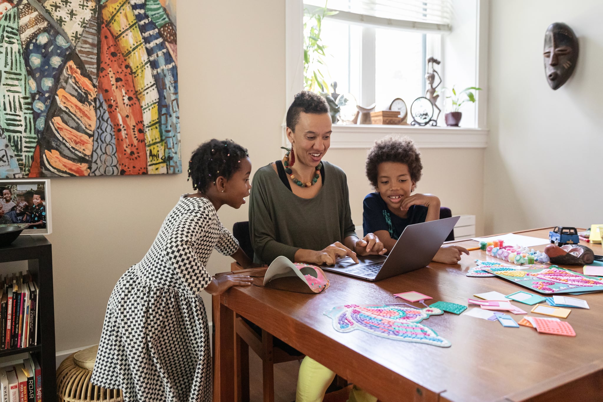 A woman sits with her son and daughter at a dining room table covered in craft materials