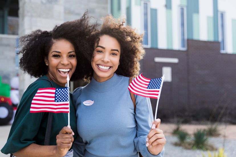 Two mid adult female friends stand together waving their American flags after voting.  They are outside the polling place.