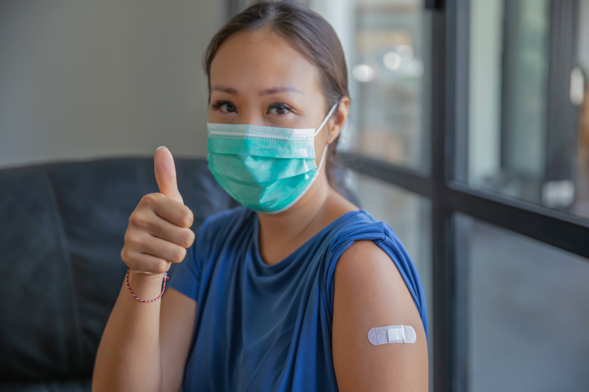 Close-up shot of proud Asian woman with protective face mask giving a thumbs up after receiving a COVID-19 vaccine at medical clinic. She's smiling behind her mask after a success vaccination.