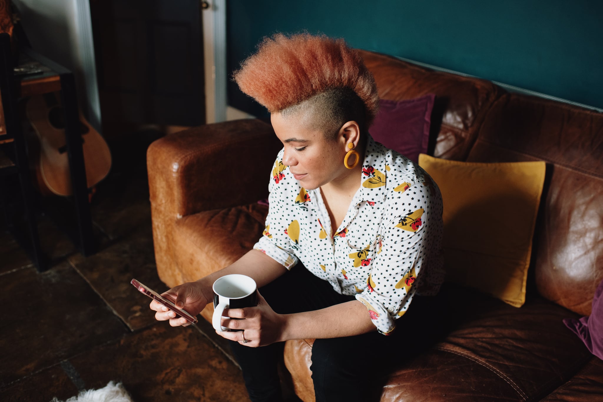 Shot from above of woman sat on sofa drinking cup of coffee and texting