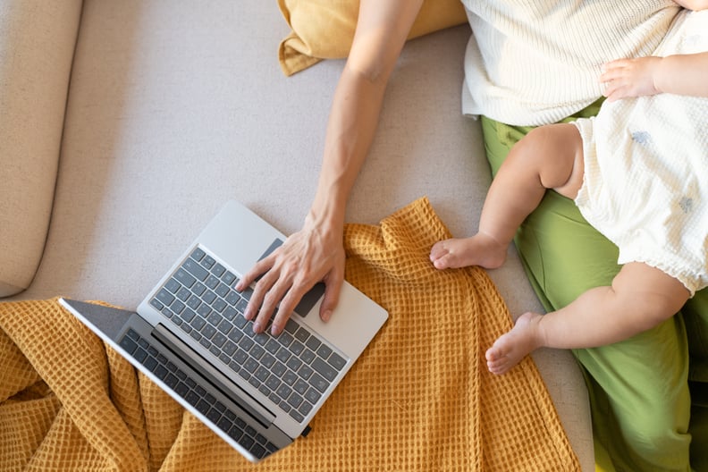 Working mother using laptop on sofa with baby sleeping in her arms