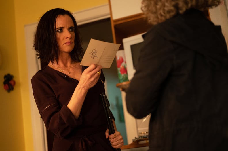 Juliette Lewis as Natalie in YELLOWJACKETS holding a postcard with the Yellowjackets rune and symbol on it