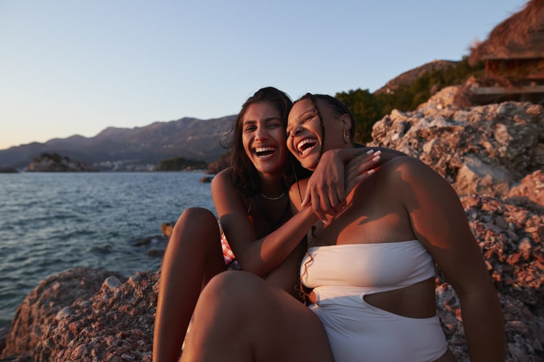 Cheerful young woman sitting on rock with arm around friend enjoying sunset together at beach