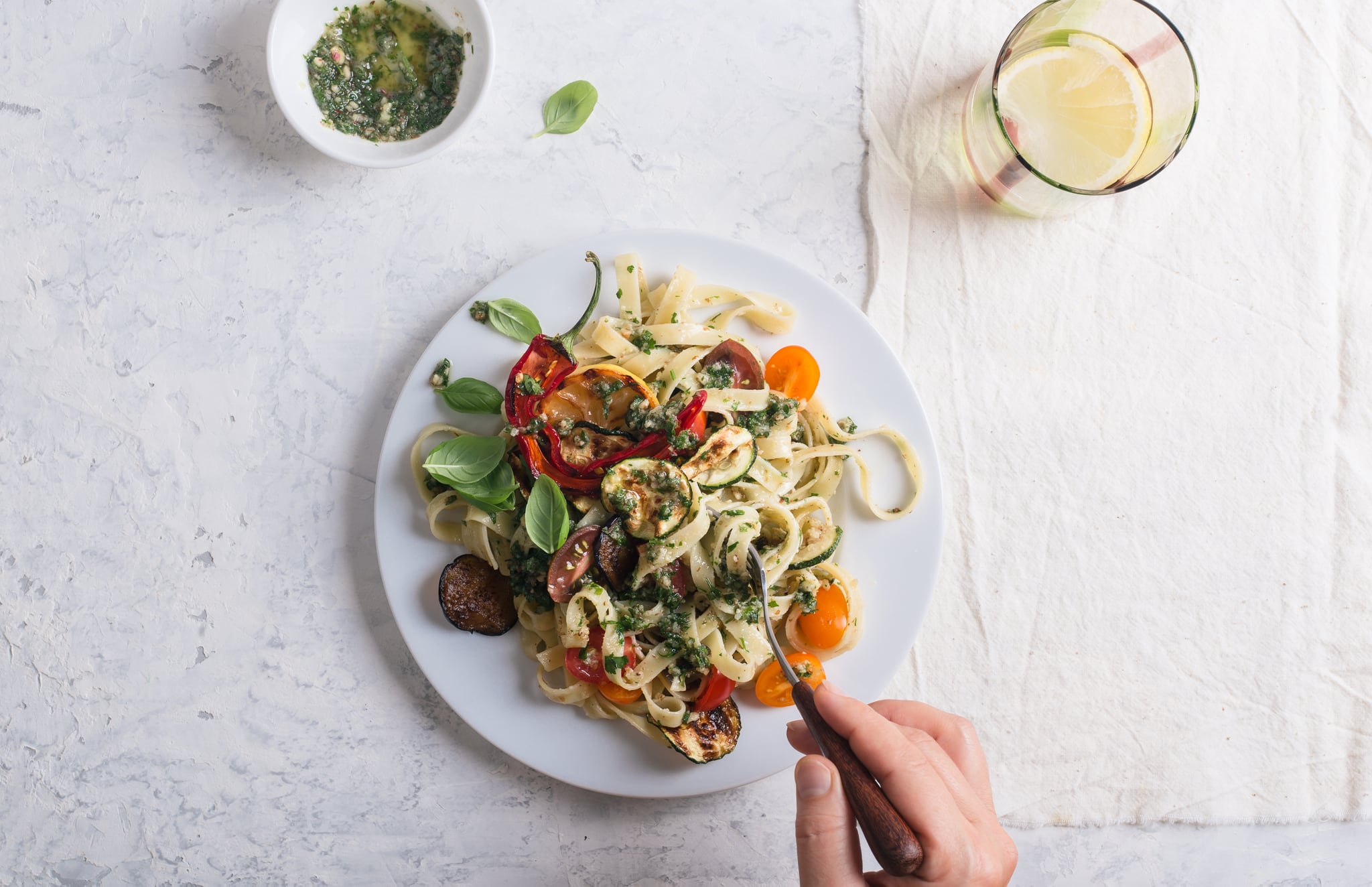 Eating Fettucine with roasted summer colorful vegetables and parsley walnut pesto, delicious vegan meal served with  fresh lemon water on rural table, top view