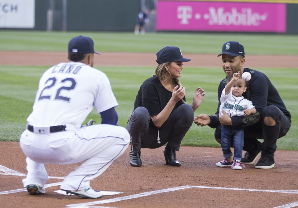 Chrissy Teigen and 1-year-old daughter Luna are currently on tour with John Legend, and on Tuesday, the brood squeezed in some quality family time at the Seattle Mariners game in Washington. While John and Chrissy looked adorable as always, it was little Luna who ended up stealing the show. With help from her mom and dad, Luna likely melted the entire crowd into a puddle when she took to the mound and threw the first pitch before the Mariners faced off against the Minnesota Twins. Naturally, Chrissy and John shared photos from their fun outing on Instagram later that evening. "Proud papa #LunasFirstTour," John captioned a snap of him holding Luna on the baseball field.
Back in 2014, Chrissy hit the field at Dodger Stadium in LA to throw out the ceremonial first pitch. Despite drinking margaritas with her dad before the big event, the model threw a nearly perfect pitch while also looking sexy in a pair of blue short shorts and knee-high socks. Clearly Chrissy passed on her baseball skills to Luna.