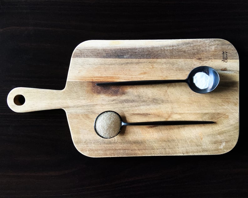 Two black spoons on a cutting board, one has sugar the other baking soda