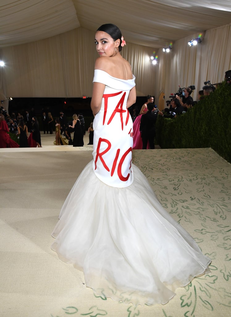 Alexandria Ocasio-Cortez made quite the debut at Monday night's Met Gala! On first glance, the American congresswoman appeared to be wearing a gorgeous off-the-shoulder white gown with a tulle train, but the second she turned around, we saw her iconic political statement: "Tax the Rich" plastered across the back of the dress in bright red lettering.
It's not lost on us that AOC chose the statement Brother Vellies look for one of fashion's most extravagant, glamorous nights. As Aurora James so perfectly said, "If you're going to come somewhere like the Met Gala and you're a congresswoman, you expect nothing less." Ocasio-Cortez completed the outfit with red heels with flowers, as well as a matching "tax the rich" bag. Ahead, see the look from the front and back.