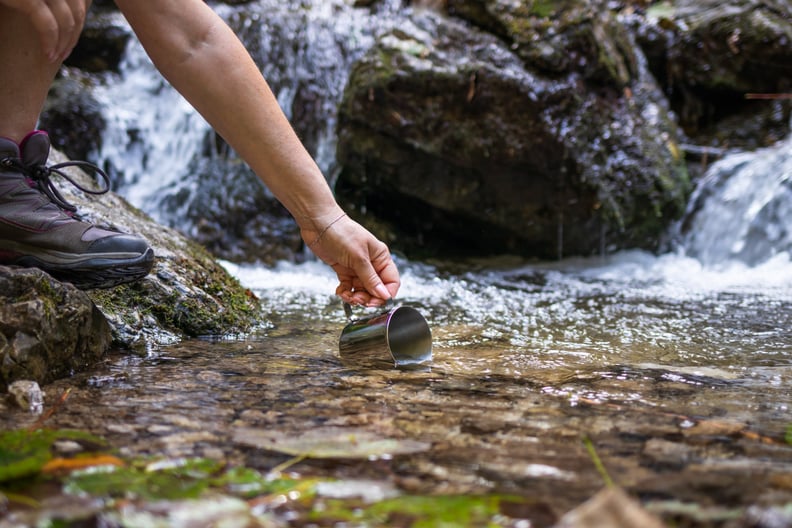 Thirsty hiker scoops fresh water into cup from stream in mountains. 