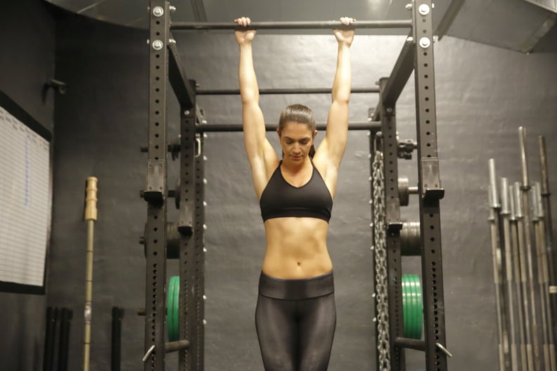 A fit, young Caucasian woman hangs from a pull up bar while doing pull ups.