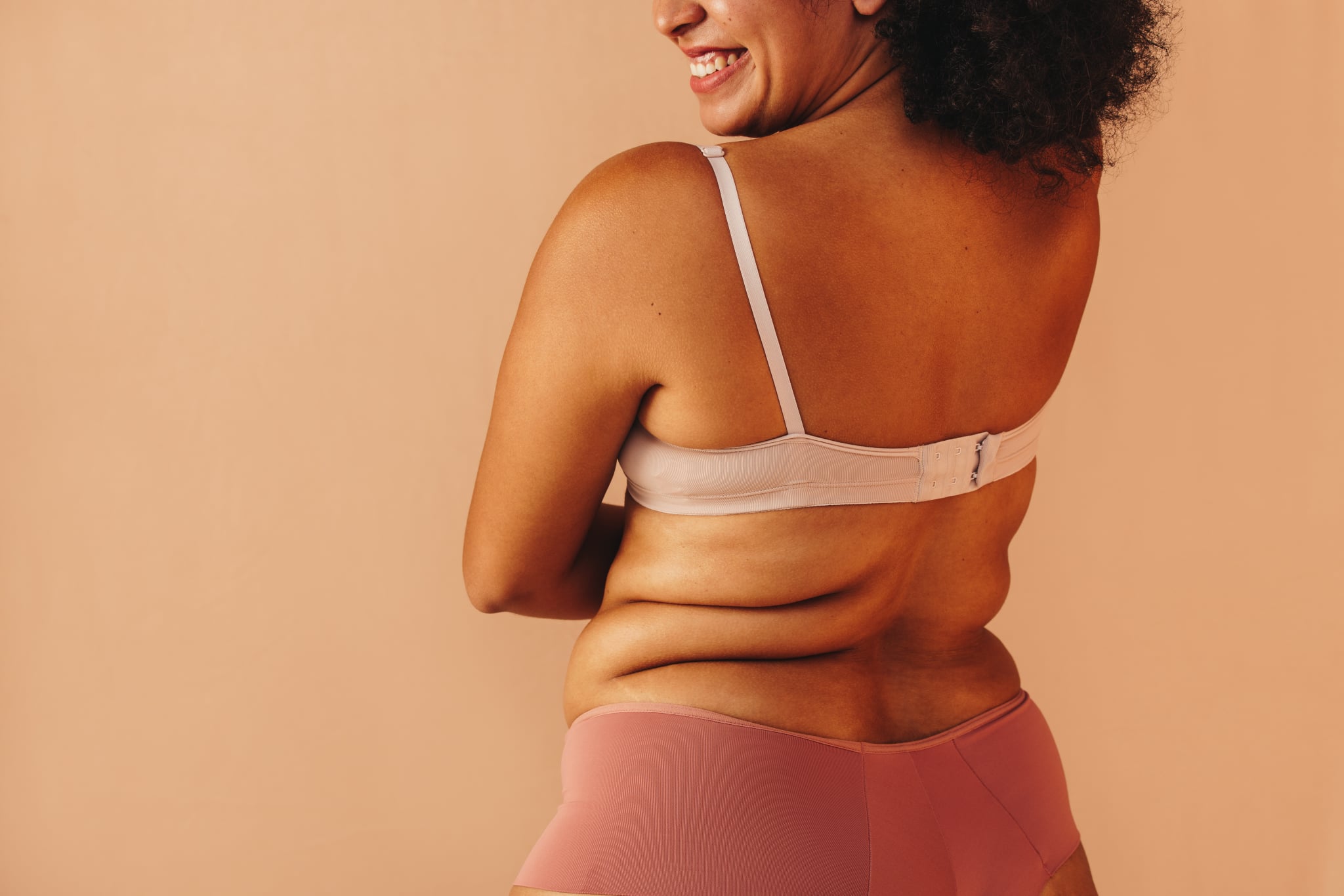 Back view of a happy curvy woman smiling cheerfully while wearing underwear in a studio. Body positive plus size woman feeling comfortable in her natural body.