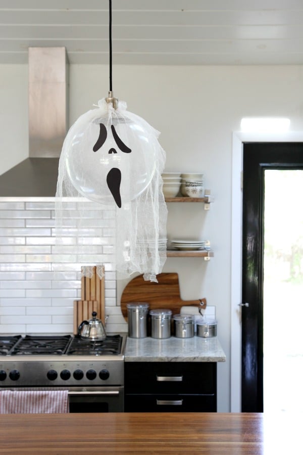 I spent $6 (coupon included) on two yards of tulle and black poster board at JoAnn’s to create ghost lights over the kitchen island. I rolled out the two yards of tulle and left it doubled over (for a layered effect), then cut it into three equal sections, one for each pendant. I cut a hole in the top of each section, slipped the tulle over the pendants, then secured it with string. I trimmed the tulle to my desired length, then “fringed” the ends by cutting ~2″ wide strips on the bottom half of each ghost. I freehanded three ghost faces and cut them out of the black poster board, then secured them to the tulle with double-sided tape.