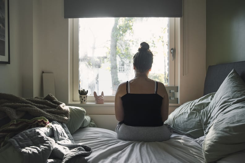 White woman in black camisole sitting on the bed looking out the window.