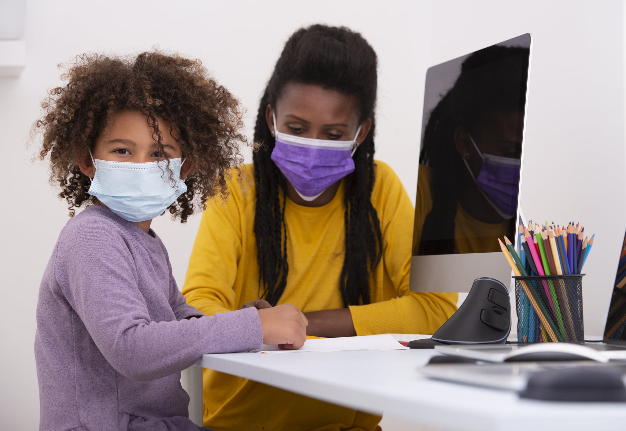 A woman teacher and school child girl wearing a cloth face-covering (face masks) due to social distancing can't be practiced. The six-year-old schoolgirl looking at the camera, studying one-on-one with the teacher and online with the rest of the class pupils, using a computer, pencils, and notebook, sitting at the table. Digital online education concept during the Coronavirus pandemic.